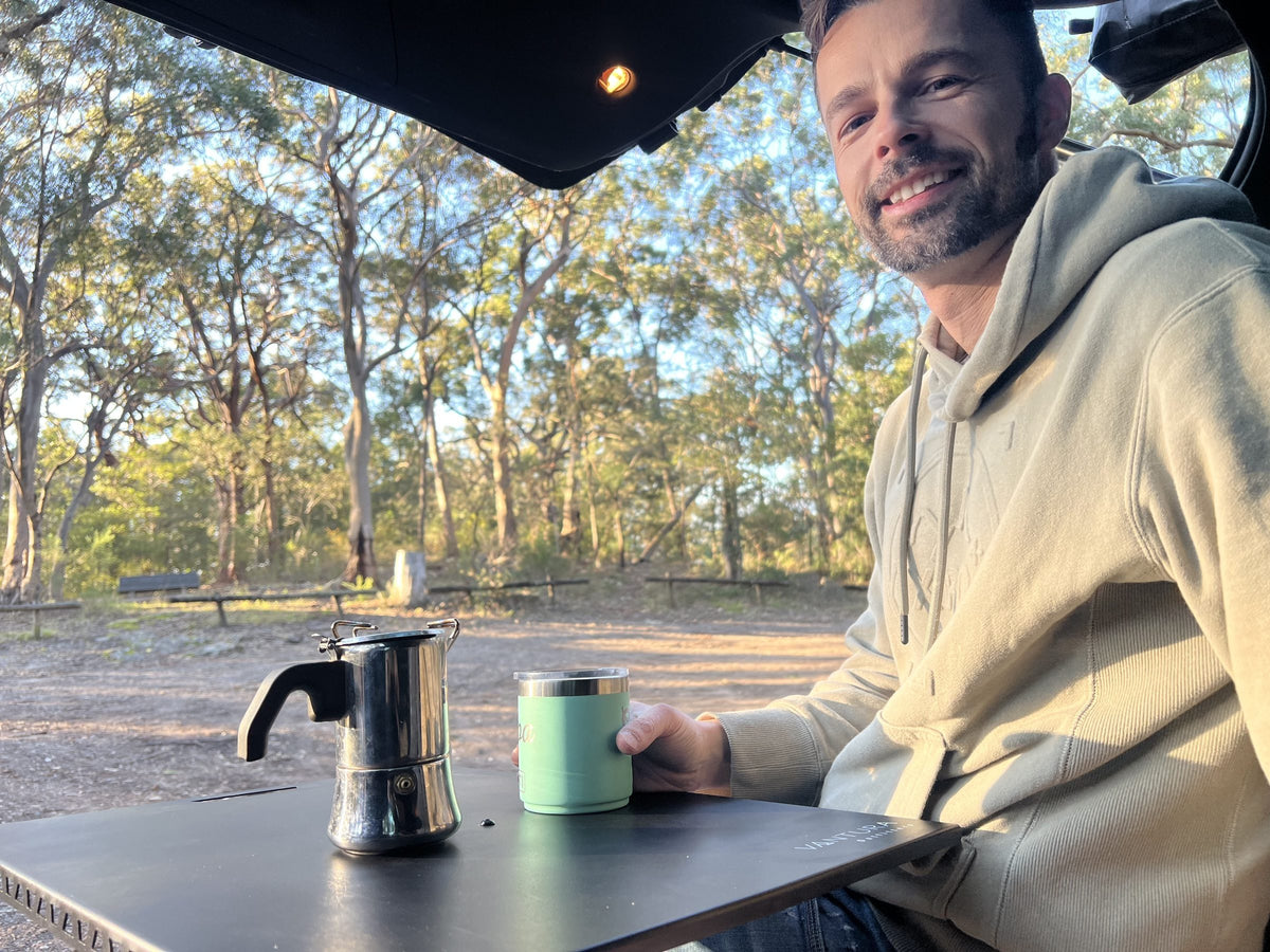 Smiling camper with coffee at the Vantura metal Bootlid table in bushland