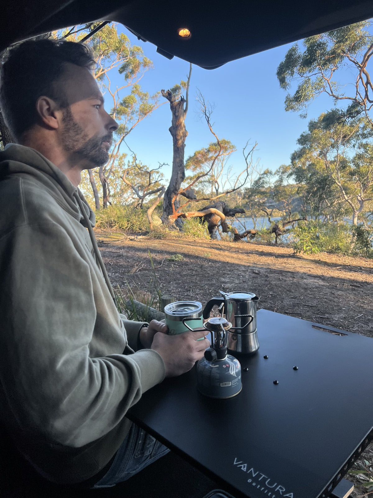 Camper making coffee at the metal Bootlid table overlooking a lake
